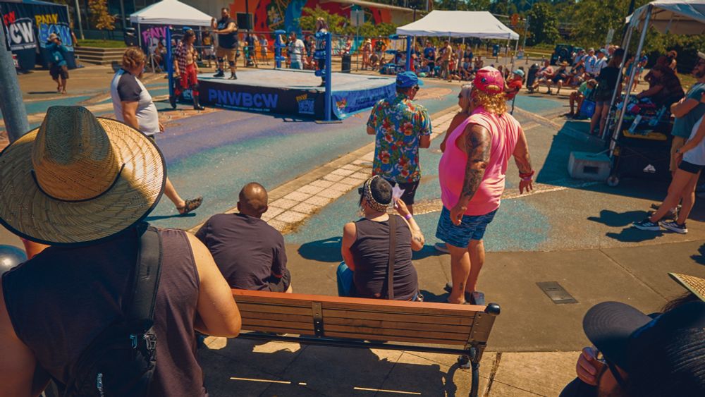 A color photo of onlookers in the crowd watching a wrestling match in the middle of the street. Caught Chris Haberman in a bright pink tank top beef caking it up.