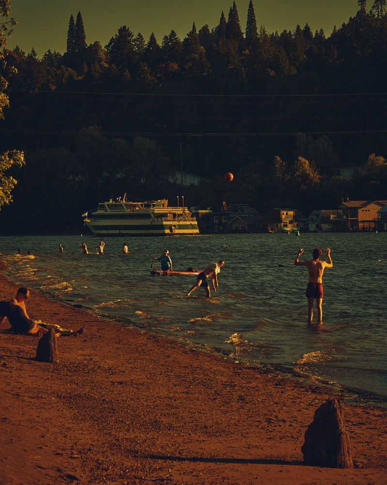 A color photo of people sitting on the beach, and playing in the water as the Portland Spirit cruises by in the background.