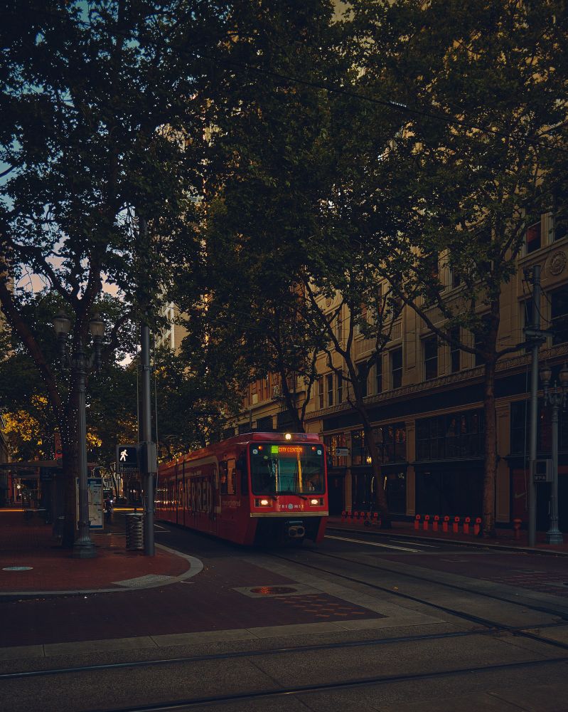 A color photo of a downtown street scene in Portland, Oregon. The MAX light rail, decked out in red, pulls up to the stop.