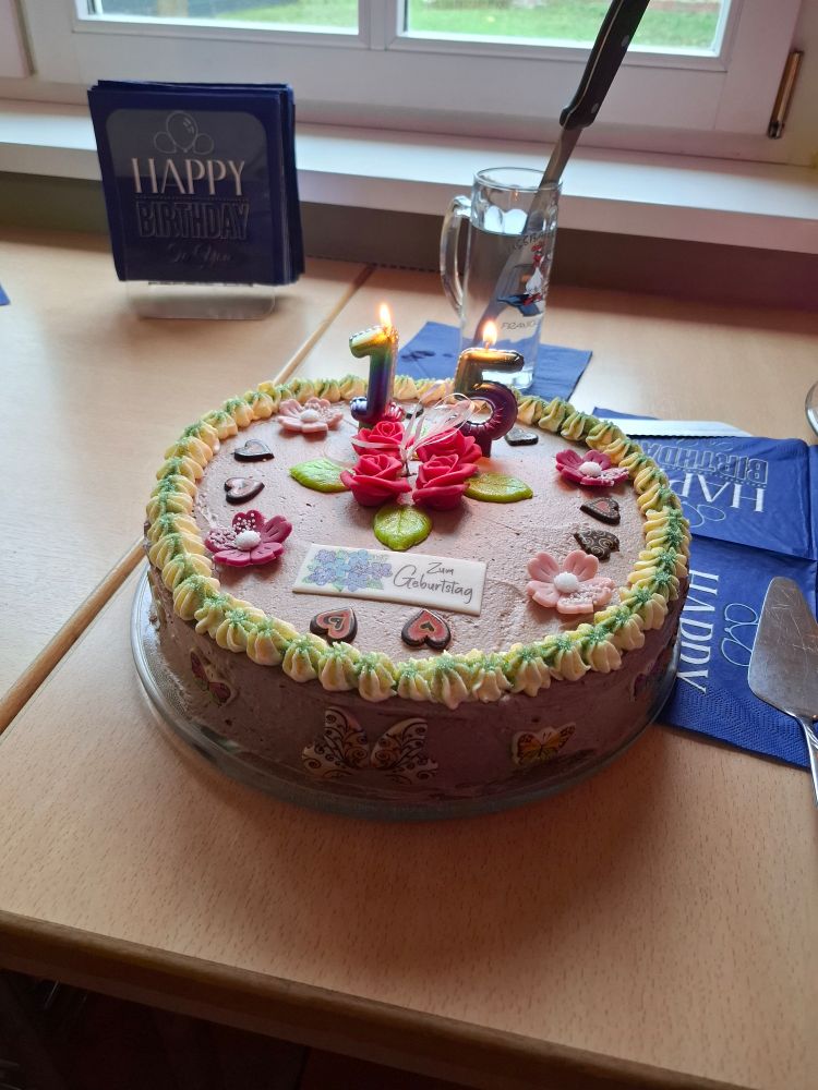 Eine Buttercremetorte,  mit Marzipanrosen, Blüten und Herzen dekoriert.  Dazu 2 angezündete Kerzen,  die eine 15 bilden. 