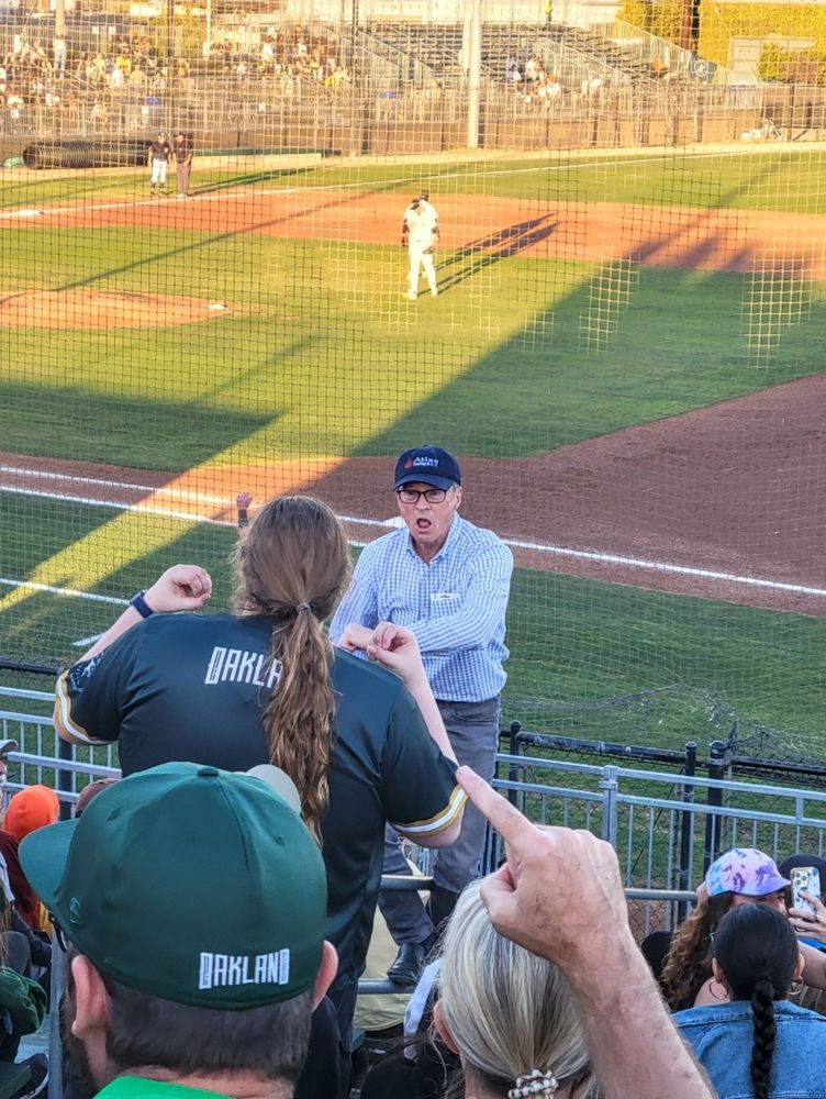 Older gentleman with a baseball cap yelling at the crowd to cheer with a woman standing up in an Oakland jersey and someone's hand in the foreground. Some baseball players are in the background. 
