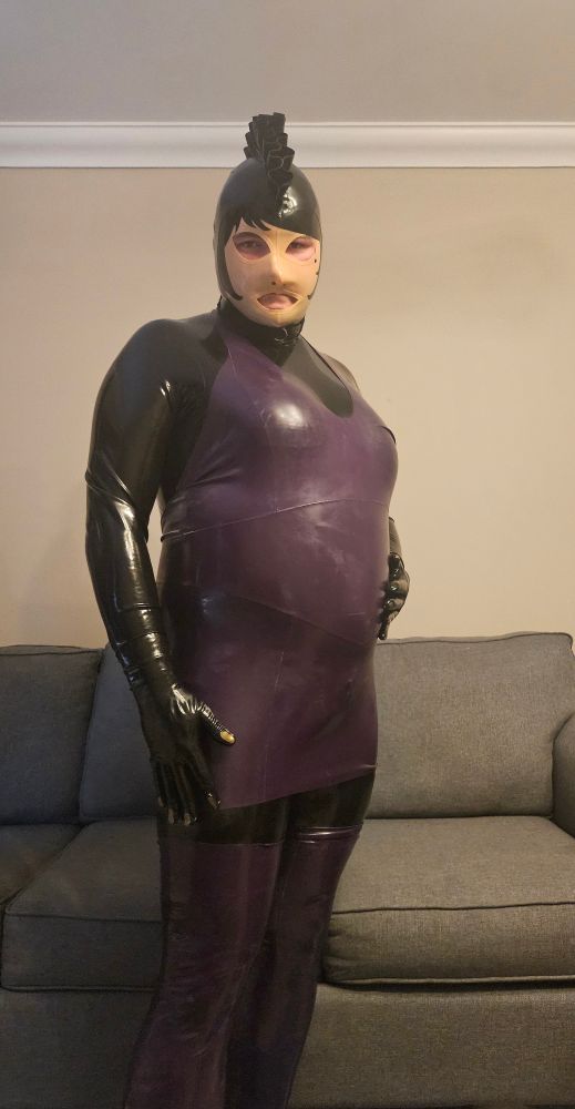 Me standing in front of my sofa wearing a black catsuit, with a purple dress, and a mannequin coloured hood with a black mohawk. Also with purple stockings on