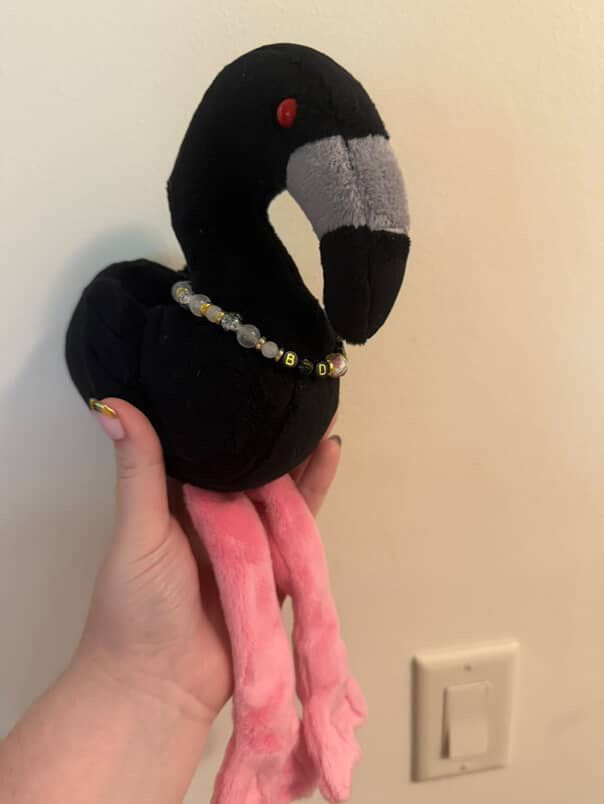 A handmade plushie of Jerry the black flamingo from Sleep Token wearing a beaded necklace that says “Baddie”. Facing to the right. 