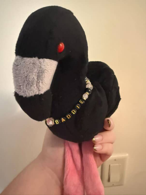 A handmade plushie of Jerry the black flamingo from Sleep Token wearing a beaded necklace that says “Baddie”
