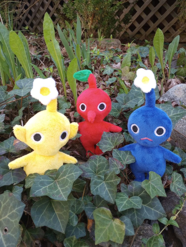 Three handmade Pikmin plushies sitting in a flower bed.  From left to right: yellow, red, and blue.