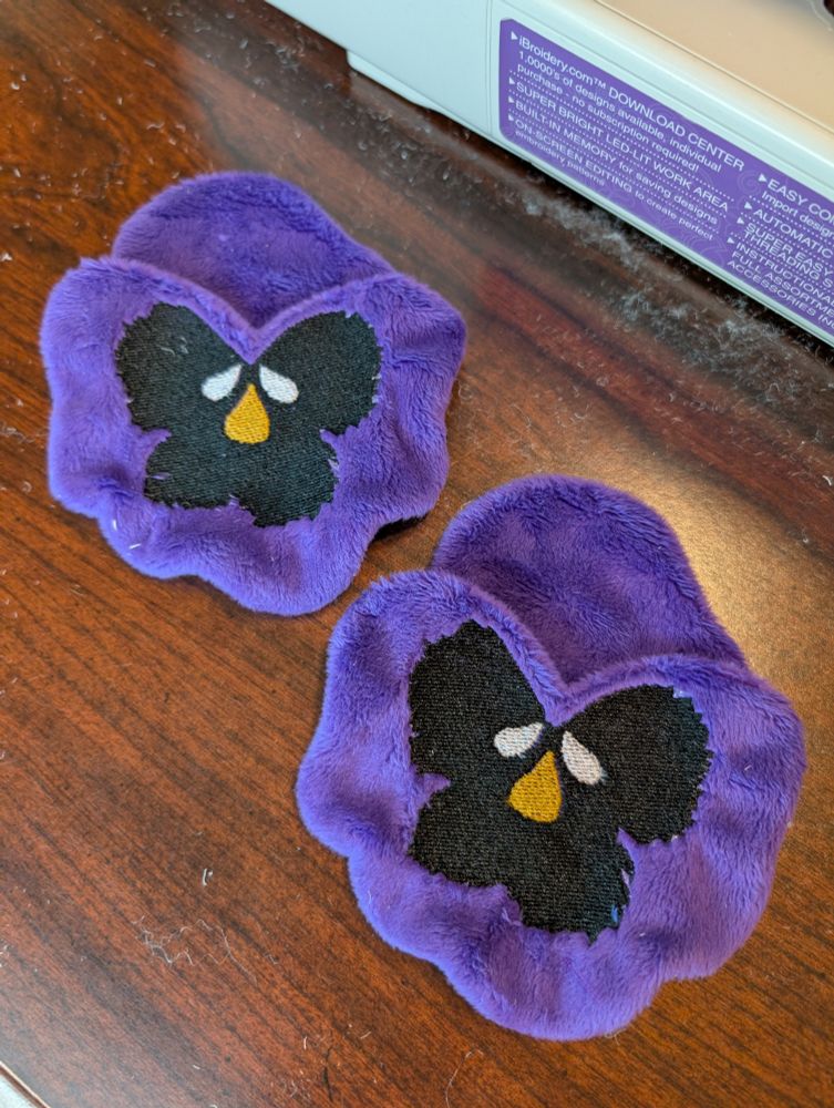 WIP of two Purple Pansy Korok plushies from the Legend of Zelda.  Shows the embroidered Pansy masks on a table.