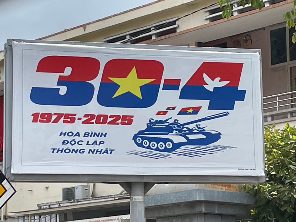 A poster with the text 30-4 1975-2025
HÒA BÌNH ĐỘC LẬP THÔNG NHẤT and a graphic of a tank, evoking the final victory of North Vietnamese forces in 1975 when a tank crashed through the gates of the Saigon presidential palace. 