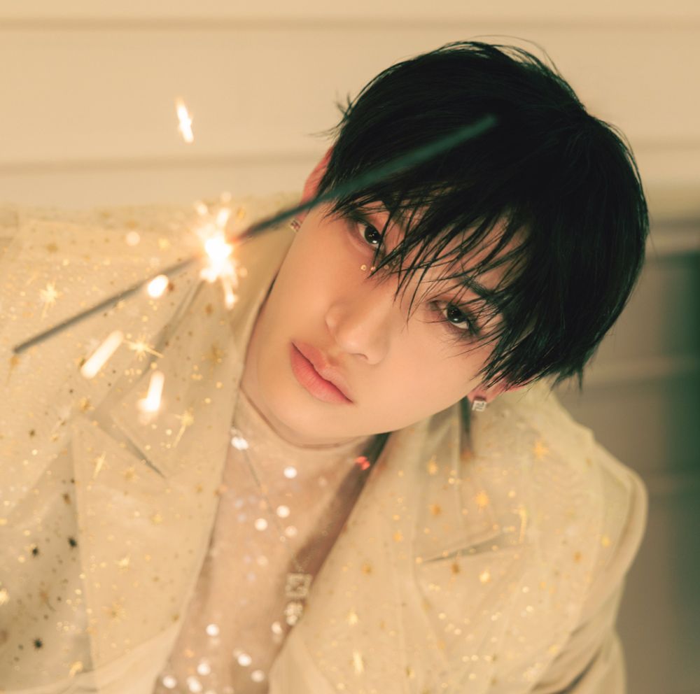 Picture shows Bang Chan wearing a sequined shirt under a tan blazer with gold flecks (not sure if painted or another technique). He is holding a sparkler.