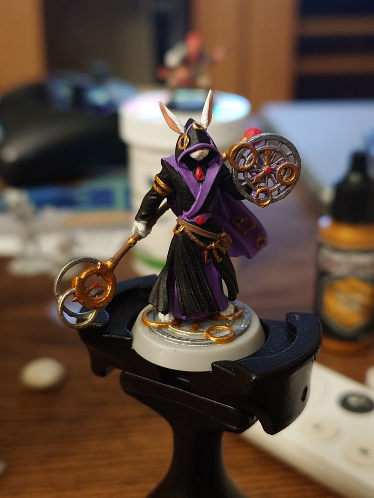 A rabbit clockwork wizard. He is wearing a black cloak, with purple trim, and gold accents. He is wielding a clockwork shield and staff.