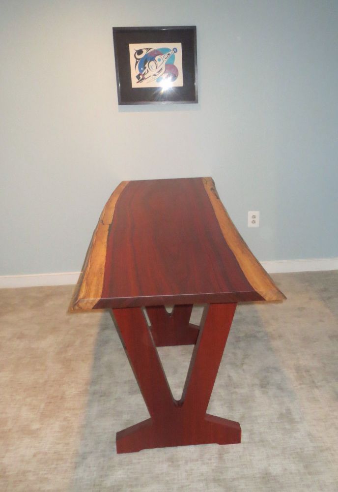 long table made from a slab with reddish heartwood and paler sapwood edges, one end is pushed up against a wall painted blue, hanging on the wall is a First Nations painting of raven in the form of a starship