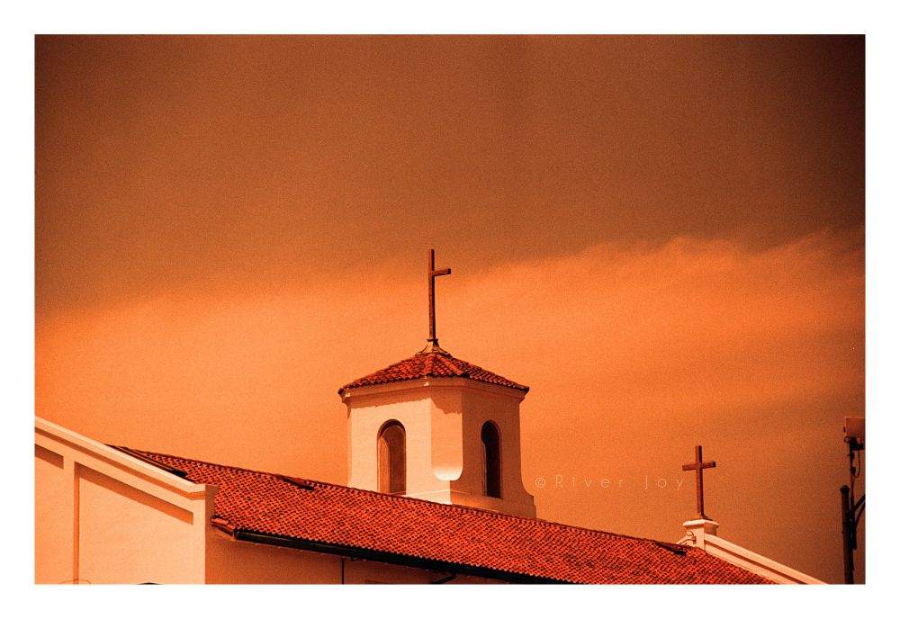 A photograph of the roof of a church, which features two crosses: the one that is higher is missing one of its arms. The photo is taken on redscale film which results in color shifts in the spectrum towards red and orange.