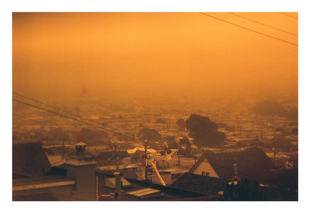 A photograph of the rooftops of Daly City on a foggy day. Some trees and bushes are visible here and there. Centered, two workers in a cherry picker are working on power lines. This image is taken on redscale film, which results in color shifts towards warmer parts of the spectrum. Everything that is not a dark shadow appears in varying tones of orange and red. 