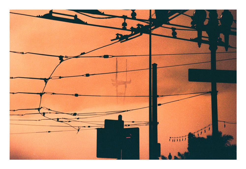 A photograph of Sutro Tower in San Francisco, California. The tower is centered in the frame, but mostly obscured by fog and clouds, leaving only the top section visible. In the foreground is a mess of cable car wires, power lines, a lamppost, street signs, and some trees all silhouetted. The shot is taken on redscale film, a process that results in everything shifting towards warmer tones on the spectrum. In this shot, the sky and clouds are a dull orange, and the only thing in the image that isn’t fully silhouetted. 