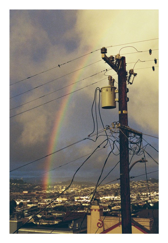 A photograph of a rainbow over a cityscape. In the foreground is a telephone pole which leans slightly to one side. Lower buildings sprawl out from the foreground towards the horizon. 

The photo is taken at golden hour, with pockets of sunlight dappling the city. 

Large clouds are arranged with sporadic holes through to blue sky. In the distance, the clouds are more stormy and grey. 

The rainbow is bright, extending from the ground and up until it disappears into the clouds. 