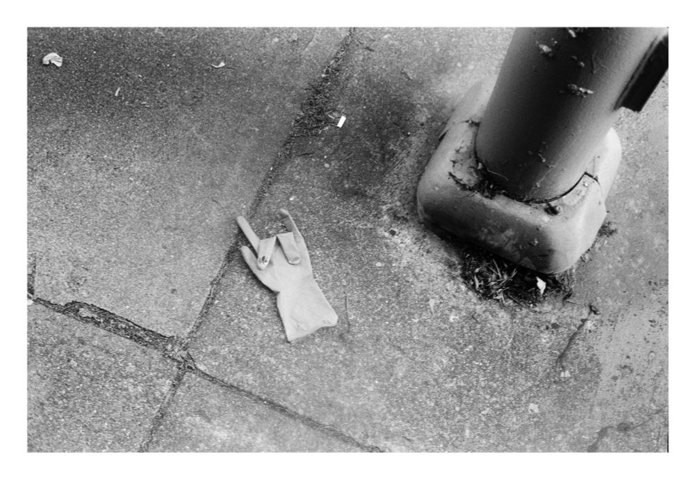 A black and white photograph of a work glove discarded on the street near the base of a street pole. The fingers of the glove are folded into the “rock on” gesture. 