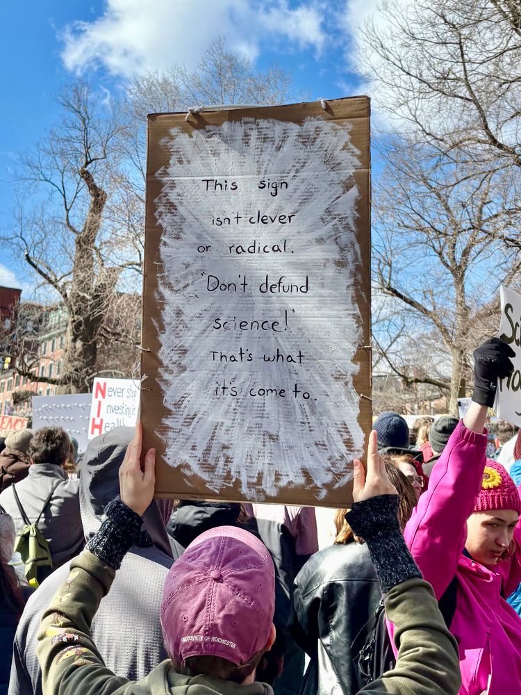 A person holds a sign saying “this sign isn’t clever or radical. Don’t defund science! That’s what it’s come to.”