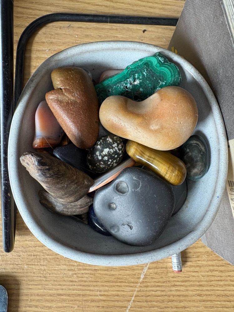 A ceramic bowl of rocks of different shapes, colors, and textures, including green malachite, tiger's eye, and a smooth lake rock with a tiny shell indentation fossil.