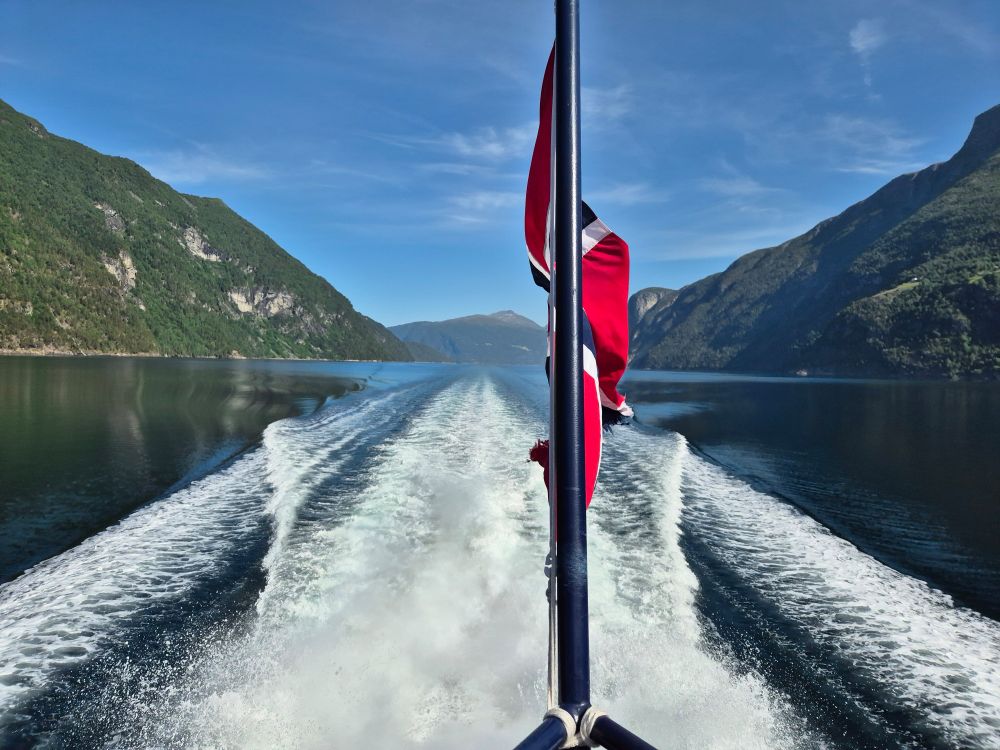Die norwegische Flagge am Heck des Schiffs teilt das Bild. Man sieht die Heckwelle des Schiffs.