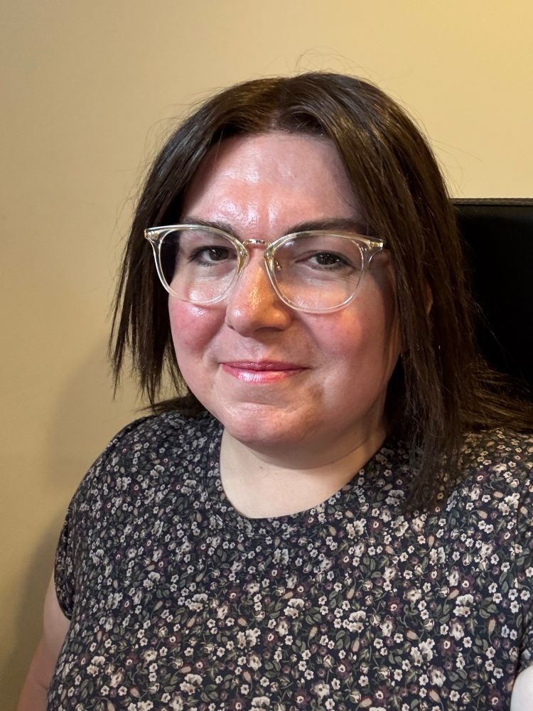 selfie of a woman smiling at the camera, wearing a floral-patterned short-sleeve top. she has shoulder-length dark brown hair, glasses with clear frames, and some light makeup (sadly with no eyeliner because my applicator dried out 😭)