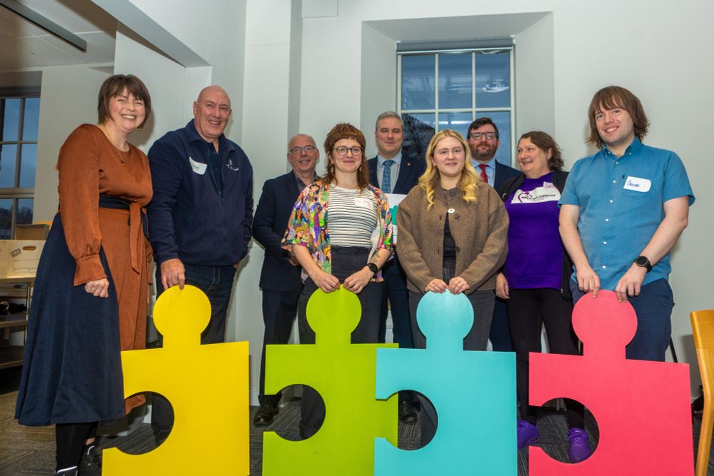 A group photograph with people smiling and standing with 4 different coloured giant jigsaw pieces (yellow, lime green, light blue and pink).