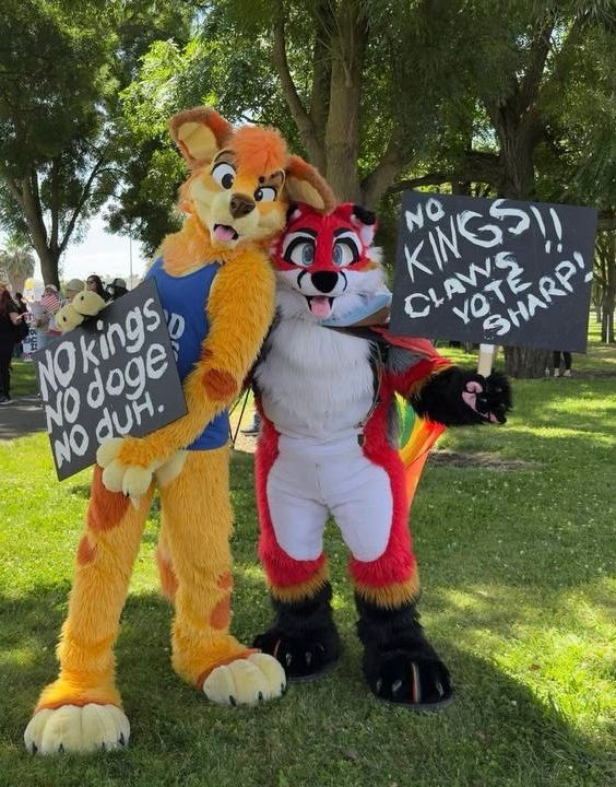 Sunny Dingo (orange yellow dingo) holding sign “no kings, no doge, no duh”, and ScoutFox (red white black fox) holding sign “no kings! Claws vote sharp!” at a No Kings event on a tree-lined path along Pacific Avenue, Stockton California
