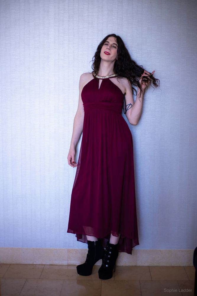 Sophie standing in a burgundy dress with a crystal collar neck and black high heel boots, passing her fingers through her long dark curly hairy