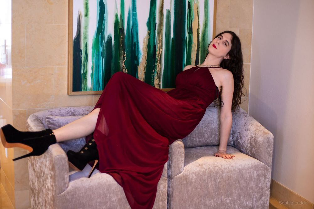 Sophie in a burgundy dress with a crystal neck and black high heels lounging on the armrests of two chairs smiling at the camera as her long wavy hair flows down behind her