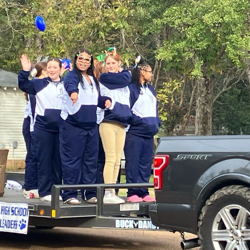 A group of high school girls are standing on a trailer being pulled by a pickup truck. The girls are wearing navy blue track suits and various Christmas hats and glasses. One girl has tossed a small blue plastic football toward the folks watching the parade. 