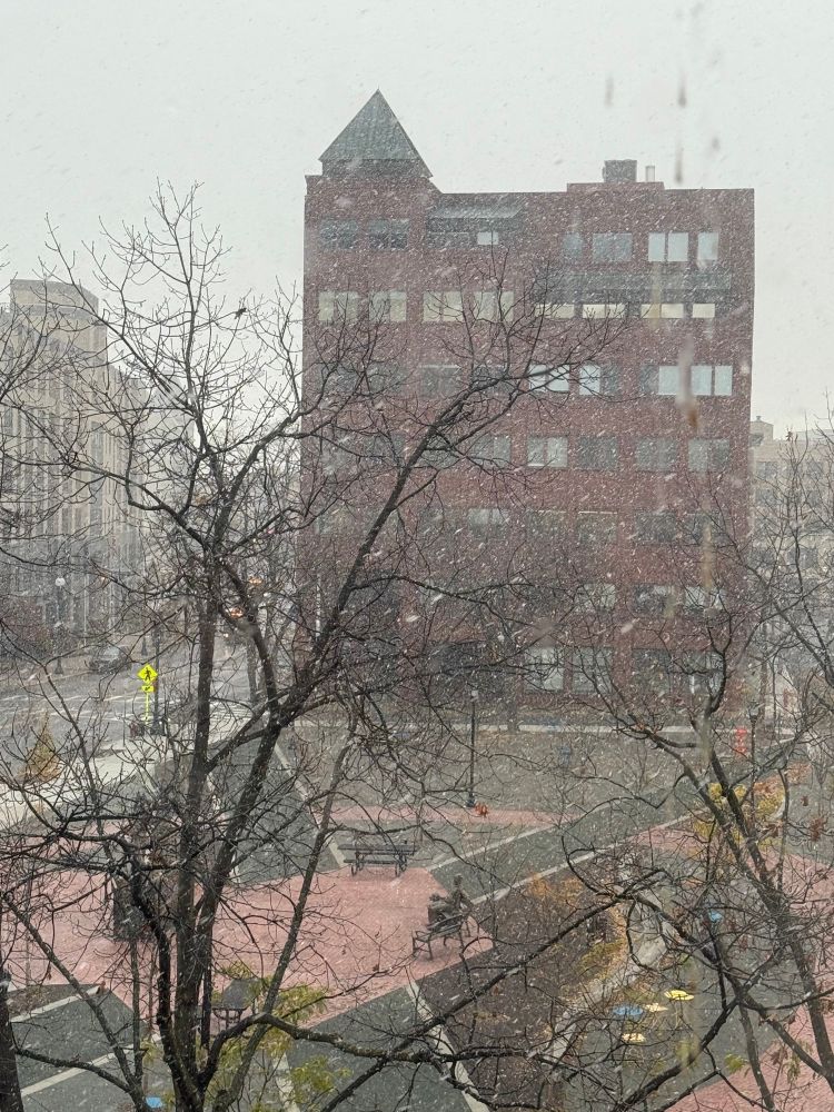 A colorful photo taken through a window of a brick & asphalt courtyard with a large, red brick building behind it. Snow flurries are seen everywhere. 