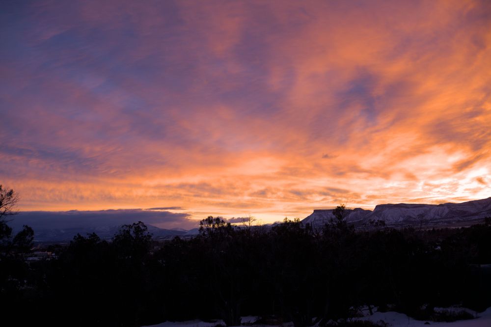 Sun just about to rise behind Mesa Verde. The ground is covered in snow, the sky is bright orange/yellow at the horizon changing to purples and blues with varying shades of orange.
The San Juan Mountains to the left are getting pummeled by a snow storm.