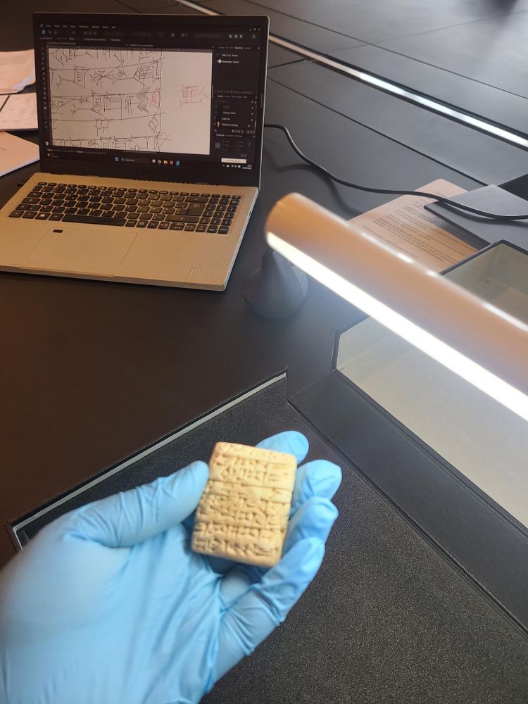Sonia Mzali tient une tablette cunéiforme dans sa main. Derrière se trouve son ordinateur sur lequel on aperçoit la copie de la tablette qu'elle a effectué.