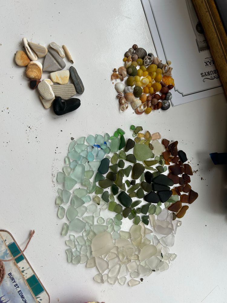 A huge pile of seaglass, organised by colour. It is mostly white, green, brown and turquoise, but there are a few rarer colours too - some tiny fragments of dark blue, some yellows, and a salmon-coloured piece. Next to it are piles of cool shells and worn pottery pieces.