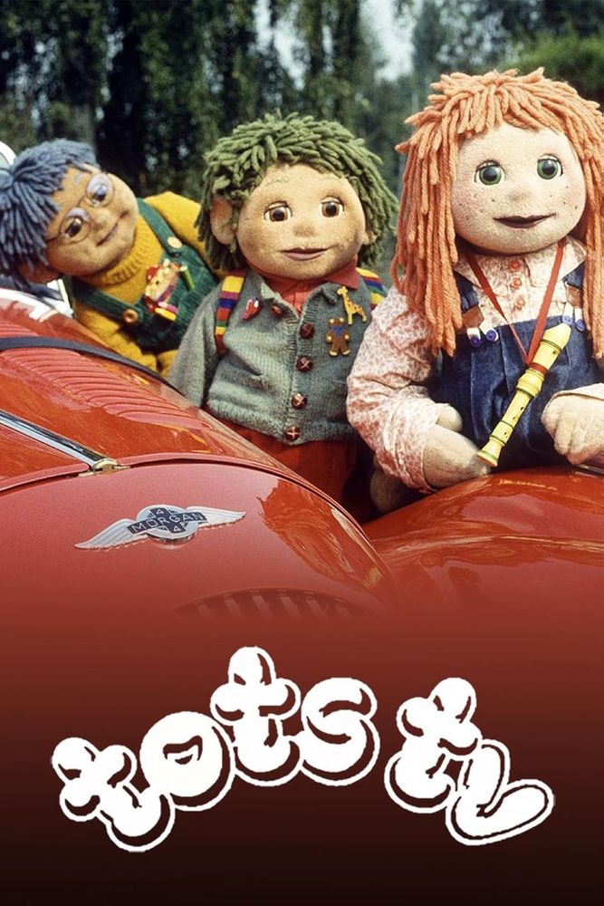 British 90s TV programme, Tots TV. Three rag dolls stand alongside a red car. One has blue hair and glasses. One has green hair and a green cardigan. The other has red hair and a red shirt.