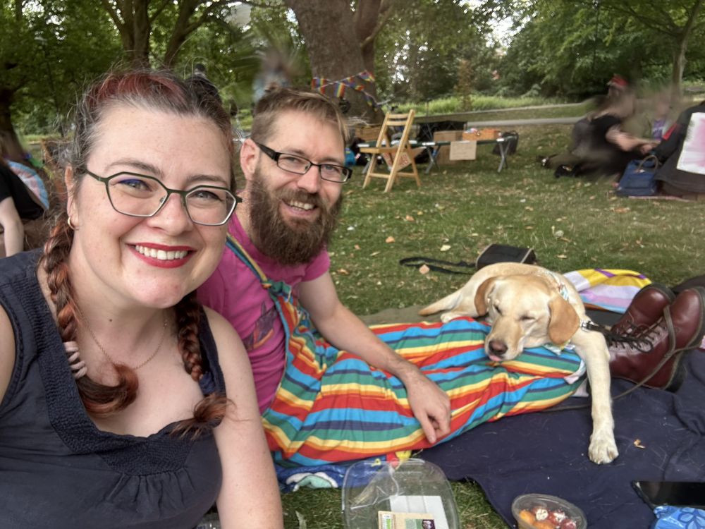 Amanda and Chris are at a picnic with their dog Lenny. Amanda has her hair in two French braids and is wearing red lipstick. Chris is wearing rainbow stripes dungarees. Lenny is a yellow labrador, and he is draped across Chris' legs. He is mid-way through licking his nose so his tongue sticks out a little. Everyone looks like they are having the best time!