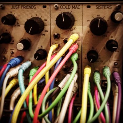 Narrow depth of field close up of the Mannequin's eurorack modules "Just Friends", "Cold Mac", and "Sisters" patched near to capacity w/ rainbow of colored cables.