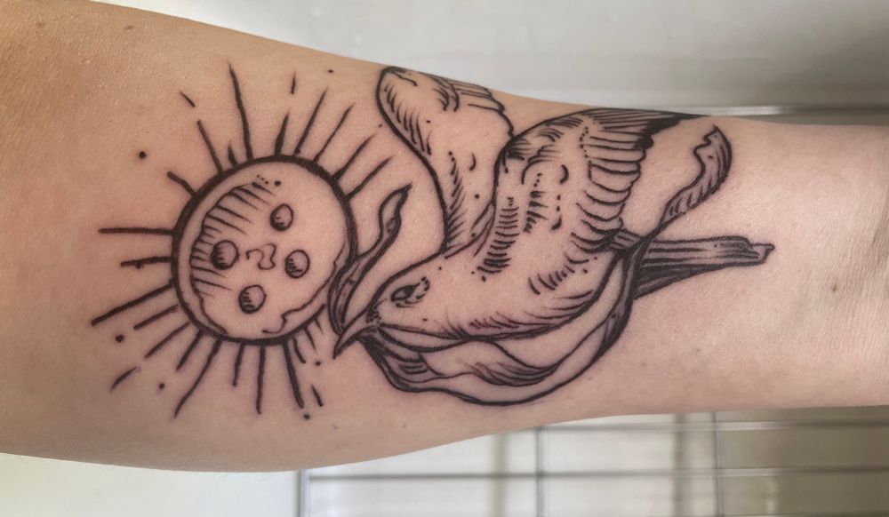 A tattoo of a seagull holding a piece of silk in his beak, flying past a button-shaped sun