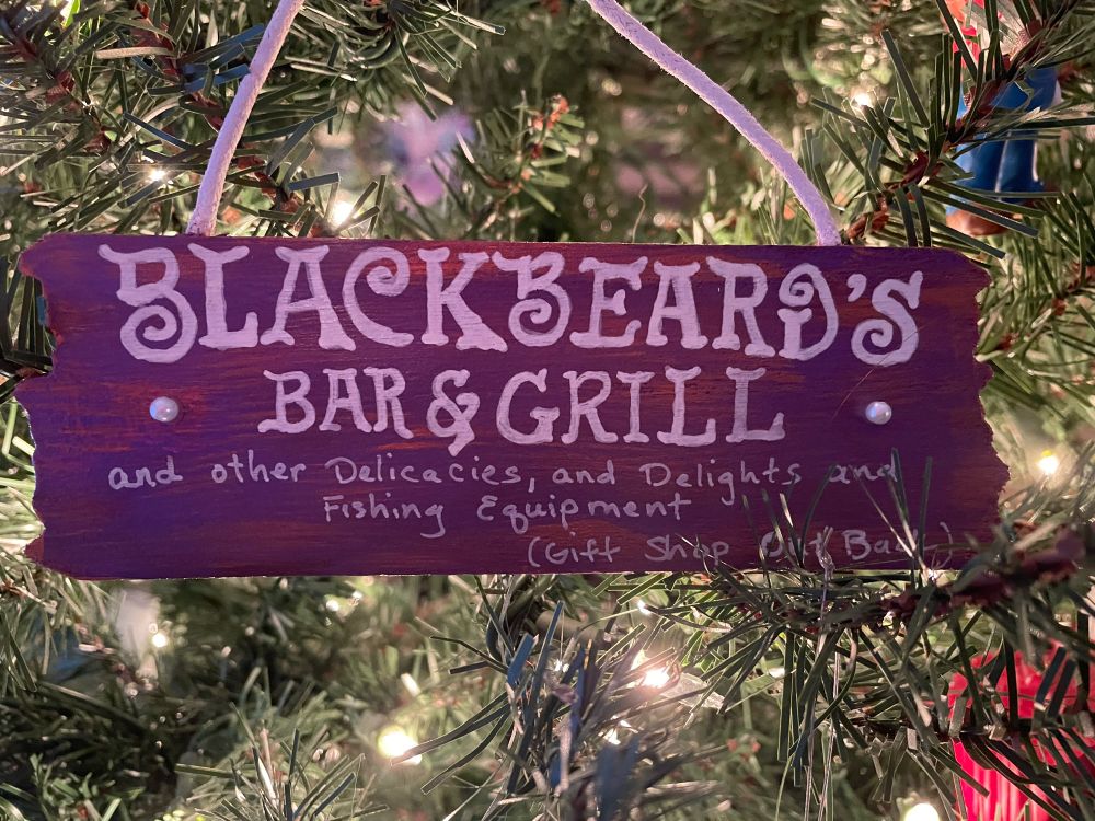 Purple/bronze painted wooden ornament that says “Blackbeard’s Bar & Grill, and other delicacies and delights, and fishing equipment…gift shop out back”