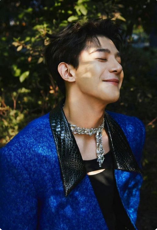 Pic of Xia Zhiguang taken from the front, in a black tank top and blue jacket wearing a wide, sparkly collar/necklace/choker, beaming widely with his eyes closed.
