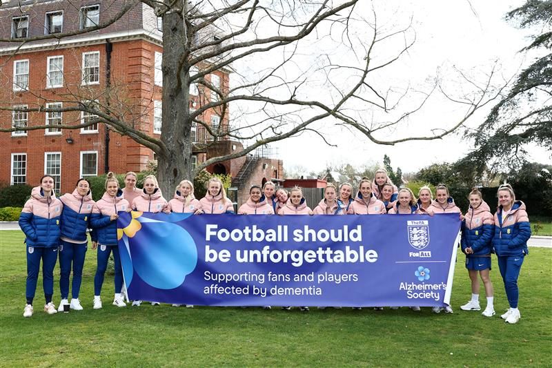 The women's England football team gathered together, standing in a line holding up a big banner. They are outside, standing on some grass in front of some trees. There are 19 of them, all wearing navy blue tracksuit bottoms and puffer jackets, the tops halves of which are salmon pink, the bottom half navy blue. They have the famous three lions crest on the chest. The banner reads: 
 
" Football should be unforgettable. Supporting fans and players affected by dementia." 