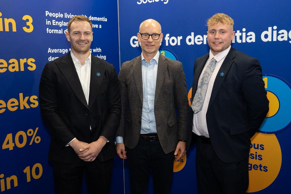 Three men pose in front of a blue backdrop with facts about dementia. They're all wearing suits and smiling.