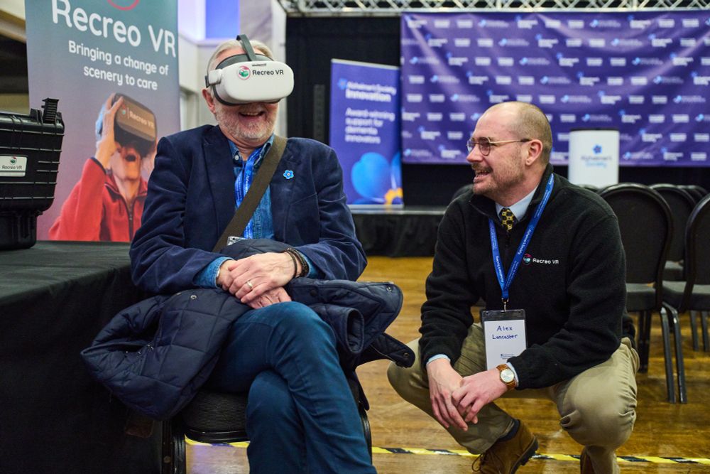 An older man in a suit sits in a chair, wearing a virtual reality headset over his eyes. Another man crouches down beside him. Both men are laughing as they discuss the experience.
