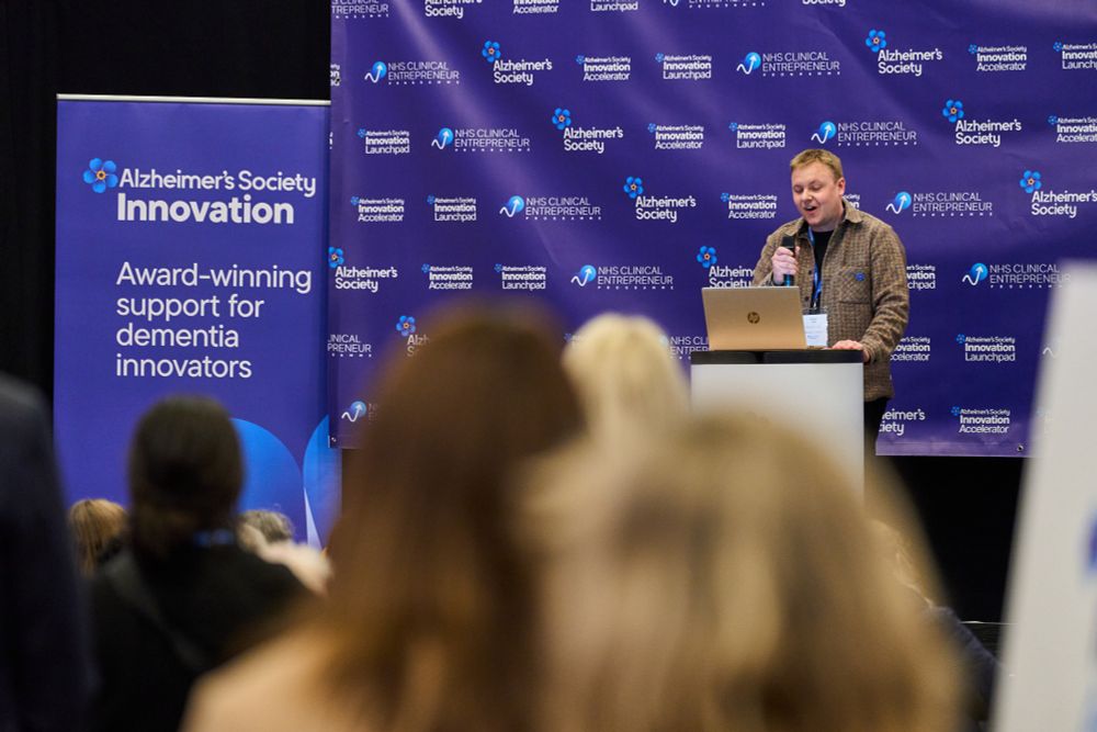 A man in his thirties with sort of red hair stands at a podium on a stage, addressing a crowd.