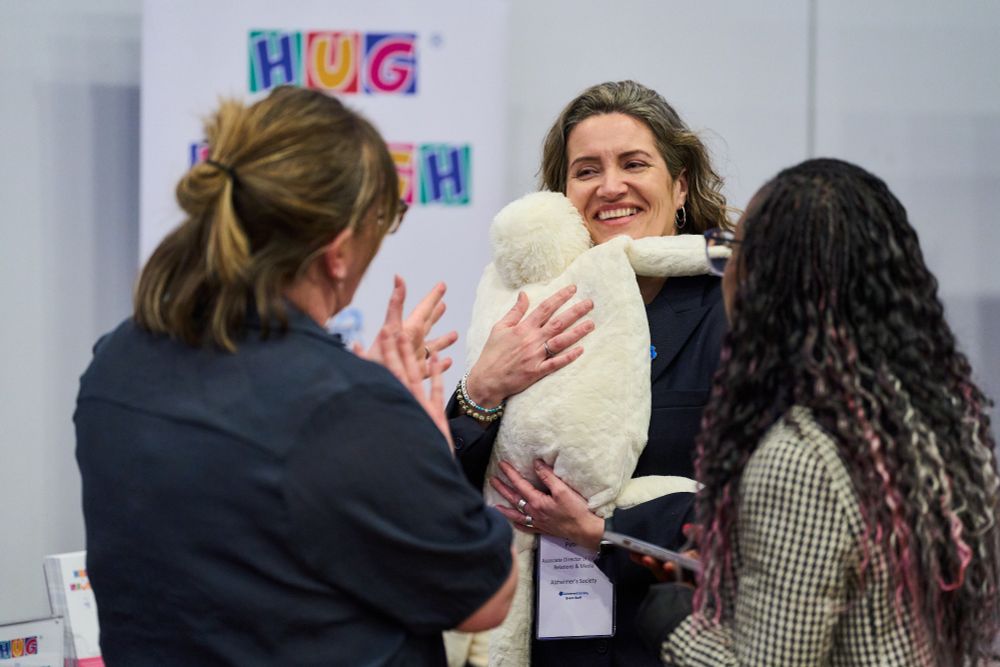 A woman in her forties with long brown hair smiles widely as she holds a toy, designed to simulate the feeling of a hug, to her chest.  