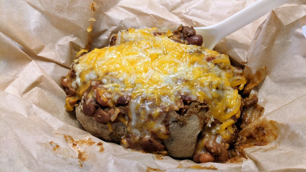 A baked potato stuffed with chili & beans and topped with melted cheese in an open brown paper wrapper