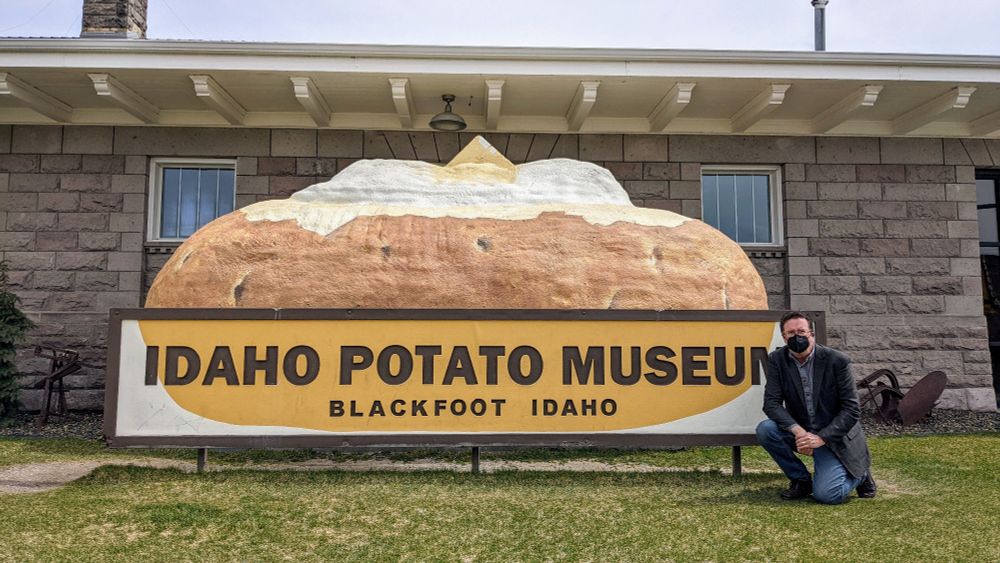 A sign shaped like a giant potato in front of a brown brick building stating that this is the Idaho Potato Museum, Blackfoot, Idaho.  There's a bespectacled man with a black KN-94 mask, jeans, and a dark sport coat, kneeling on one knee next to the sign on the right side of the photo.  It's a grey and overcast day.