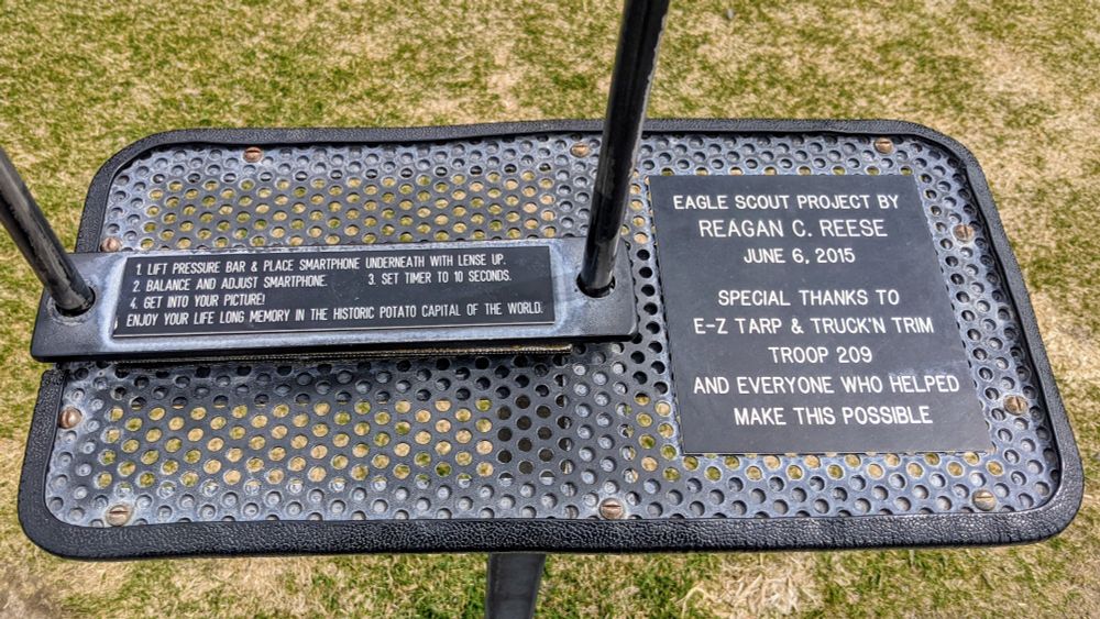 A black metal platform on a grassy lawn in muted sunlight; view is looking down obliquely at the top of the platform.  A small sign on the left side of the platform reads, "1. Lift pressure bar & place smartphone underneath with lense (sic) up.  2. Balance and adjust smartphone.  3. Set timer to 10 seconds.  4. Get into your picture!  Enjoy your life long memory in the historic potato capital of the world."  Another sign on the right side of the platform reads, "Eagle Scout project by Reagan C. Reese, June 6, 2015" and "Special thanks to E-Z Tarp & Truck'n Trim, Troop 209, and everyone who helped make this possible."
