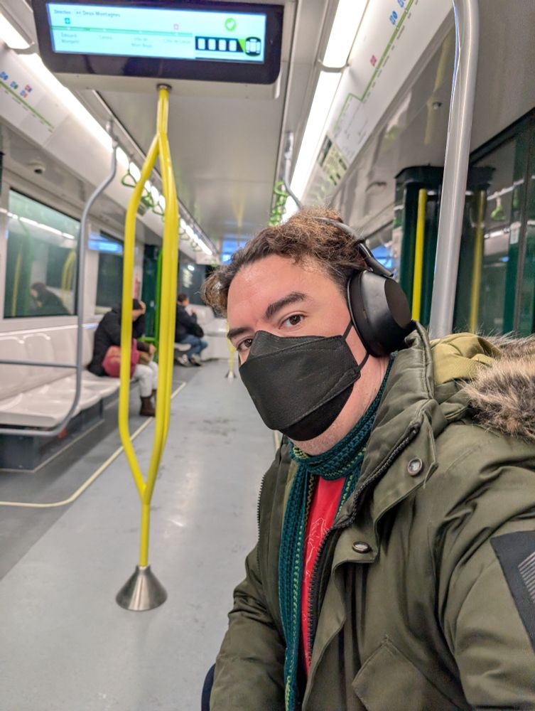 White Man in a dark green winter coat with a black face mask and black headphones. On a train with only a handful of other people.