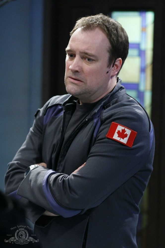 Dr Rodney McKay from Stargate Atlantis 
Played by David Hewlett

A white man with receding brown hair wearing  a gray/blue jacket. He has a shoulder patch of the Canadian Flag.