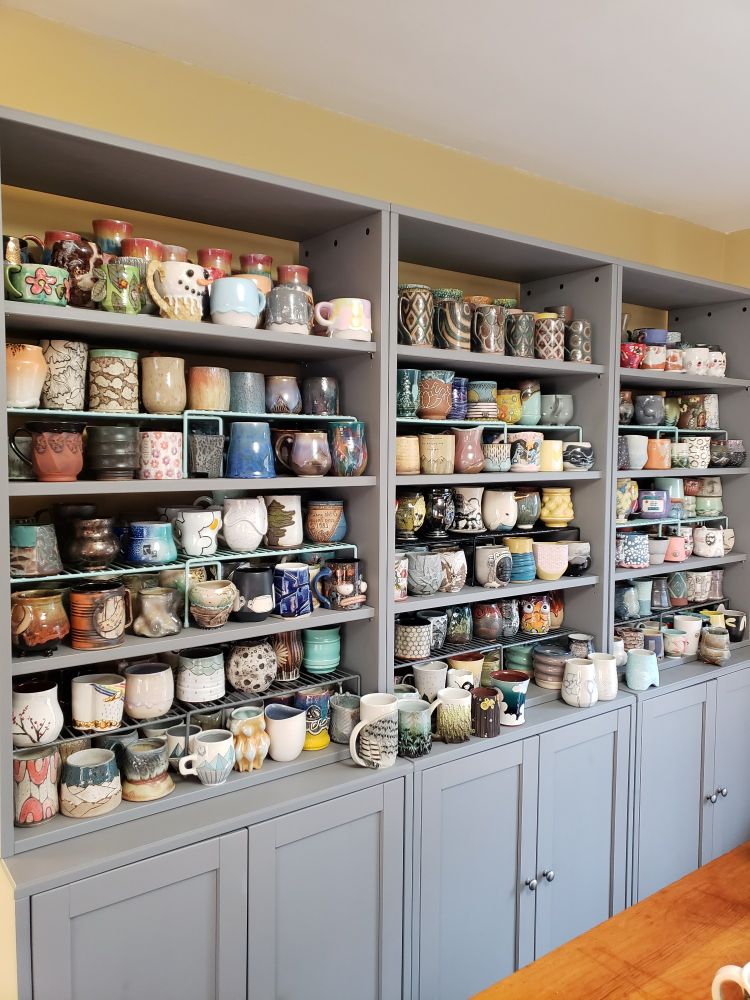Three grey cabinets are lined up with upper shelves full of hand-made, cermaic mugs and cups. There are so many shapes, sizes, and colors that it's impossible to describe adequately.