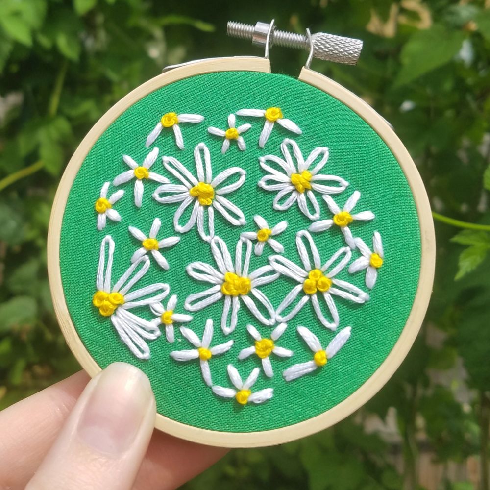 Hand holding a tiny embroidery hoop. Bright green fabric fills the hoop, with the fabric covered in tiny hand embroidered daisies in various sizes. White and yellow embroidery thread makes up the daisies. 